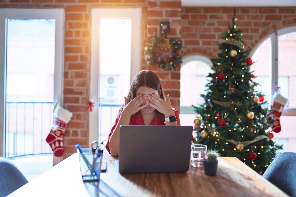 Woman under holiday stress, with hands covering eyes, sitting in front of laptop looking frustrated, with Christmas tree in background.