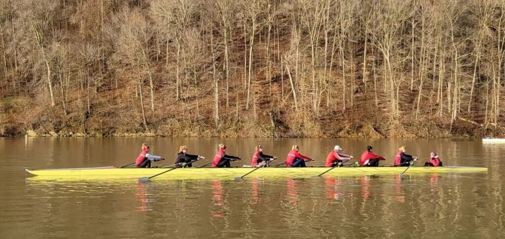 A women's rowing crew rowing along a river, against a backdrop of wintery trees.